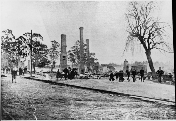A black and white image of a building ruined by fire, only three brick chimneys remain standing.